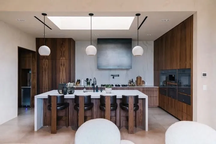 Symmetrical walnut kitchen with a white waterfall island, four stools, globe pendants and a skylight