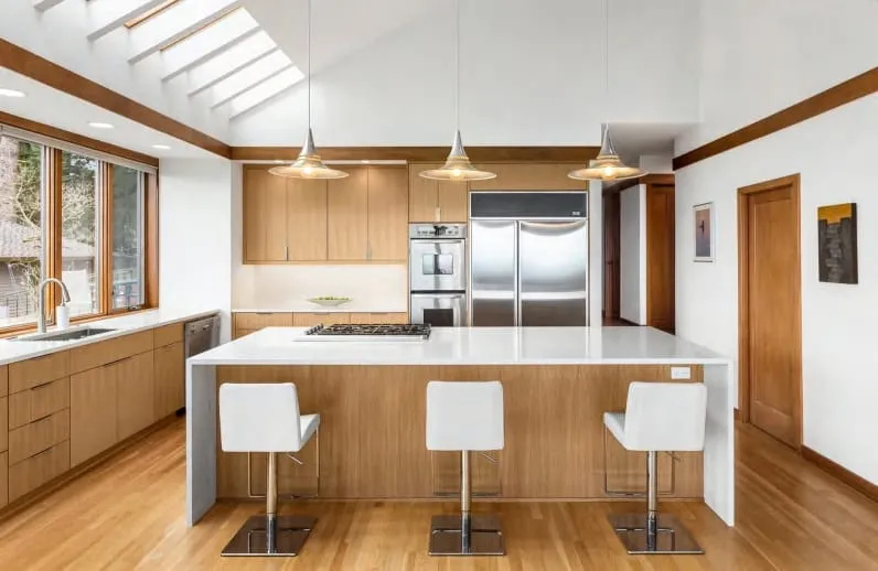 Front view of a white island with a cooktop under three pendants, with stainless refrigerator and wall ovens behind