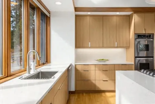 Oak cabinets around a stainless sink by tall wood-framed windows, with double wall ovens