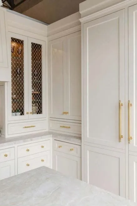 Tall greige cabinet with long brass pulls next to a cabinet with a wire grille door insert