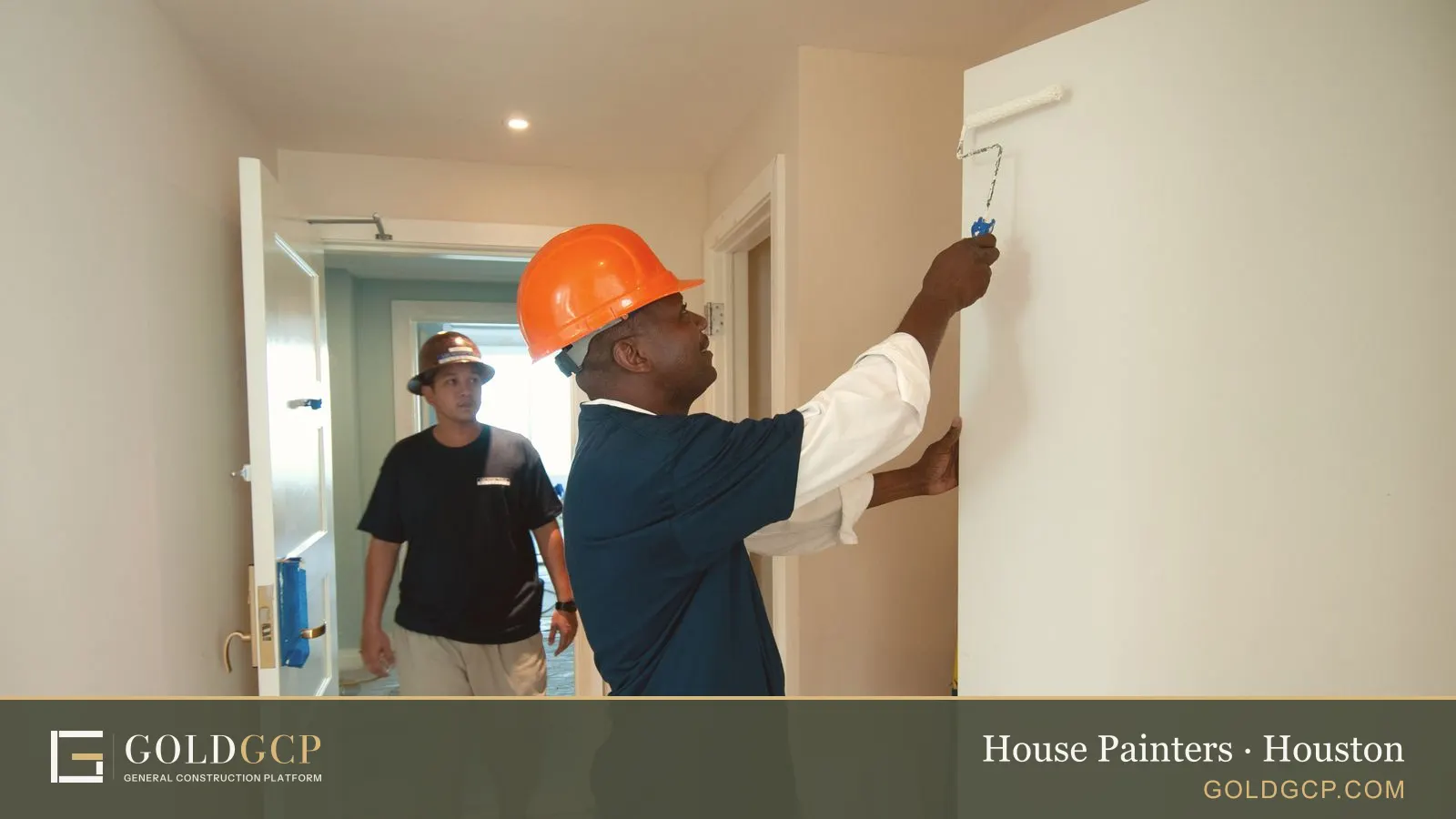 A painter rolling paint onto an interior wall while a coworker walks the hallway