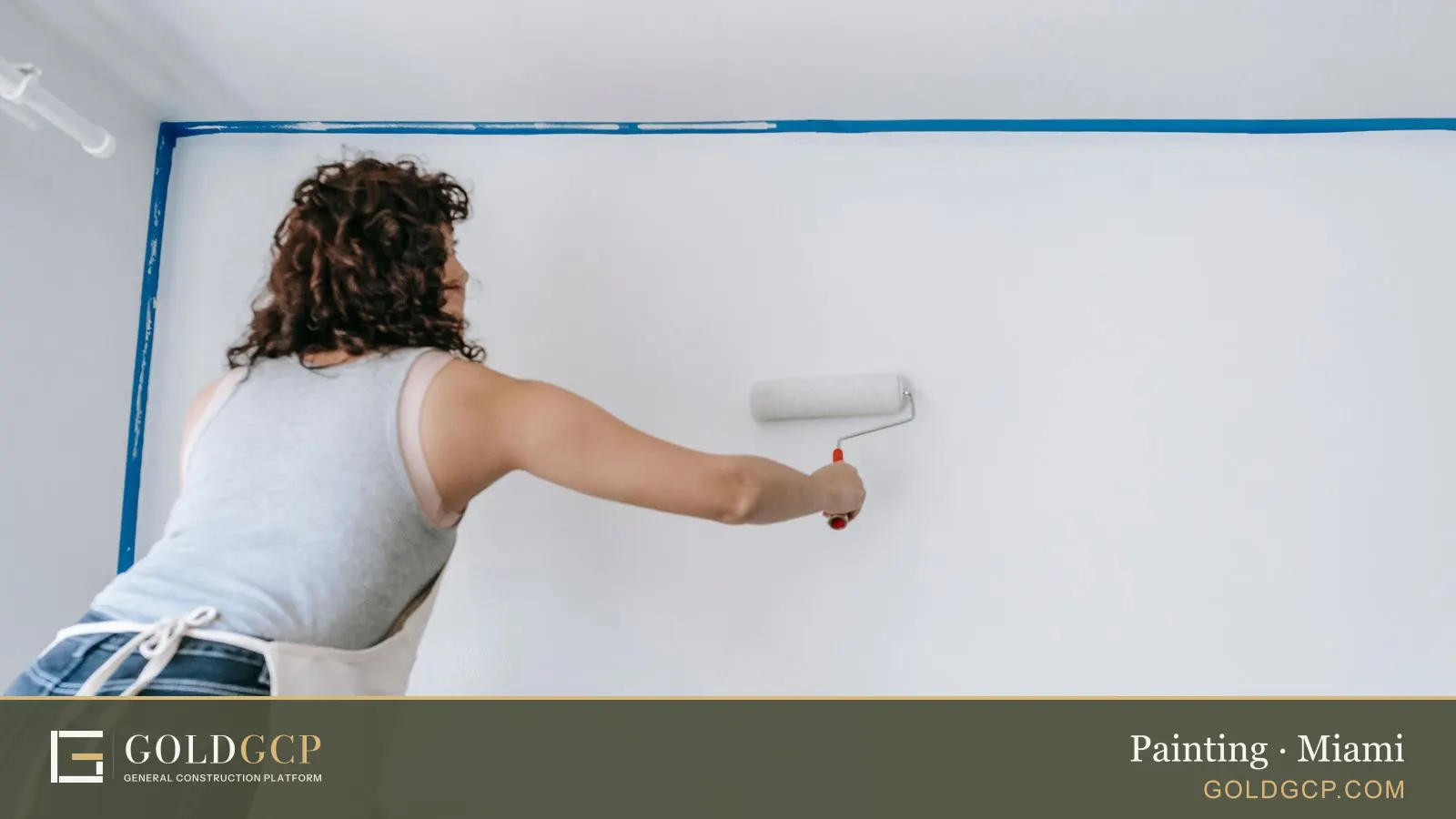 A painter rolls white paint onto a wall edged with blue painter's tape